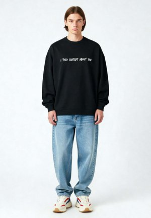 Zwarte sweatshirt met witte tekst "IK HEET CHATGPT OVER JOU," gecombineerd met lichtblauwe jeans en meerkleurige sneakers. Soepele stofstructuur.