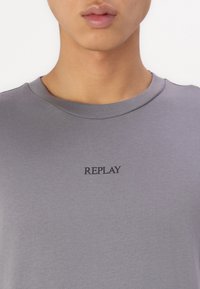 Grijze katoenen T-shirt met een ronde hals en korte mouwen; heeft een zwart "REPLAY" logo gecentreerd op de borst. Gladde textuur.