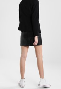 Personne portant un blazer noir et une mini-jupe en cuir avec des baskets blanches montantes, se tenant devant un fond clair.