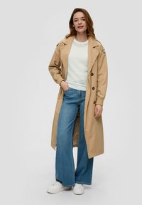 Jasno beżowy płaszcz trench z paskiem w talii, nałożony na teksturowany biały sweter, w połączeniu z szerokimi niebieskimi dżinsami i białymi trampkami.