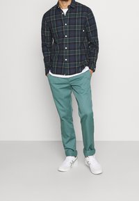 Hombre con camisa de botones a cuadros en verde oscuro, pantalones enrollados en azul claro, zapatillas blancas, de pie con las manos en los bolsillos contra un fondo liso.