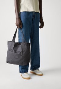 Persona che indossa jeans blu, top crema, sneakers bianche, e tiene una borsa a tracolla grigia scura con la scritta "ALLSAINTS LONDON".