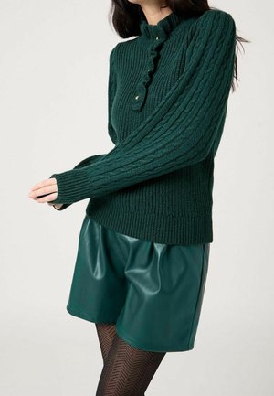 Femme portant un pull en maille torsadée vert foncé avec des boutons et un short en cuir vert assorti, associé à des collants noirs à motifs.