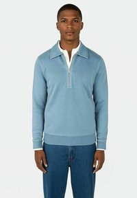 Junger Mann in einem hellblauen Pullover mit Viertelreißverschluss über einem weißen Hemd und blauen Jeans, der vor einem schlichten weißen Hintergrund steht.