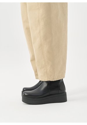 Person iført beige bukser og sorte Chelsea-støvler med plateau, stående på en hvid baggrund.