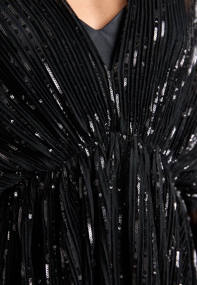 Femme portant une robe noire avec des rayures verticales de sequins brillants et un décolleté en V profond sur des vêtements sombres.
