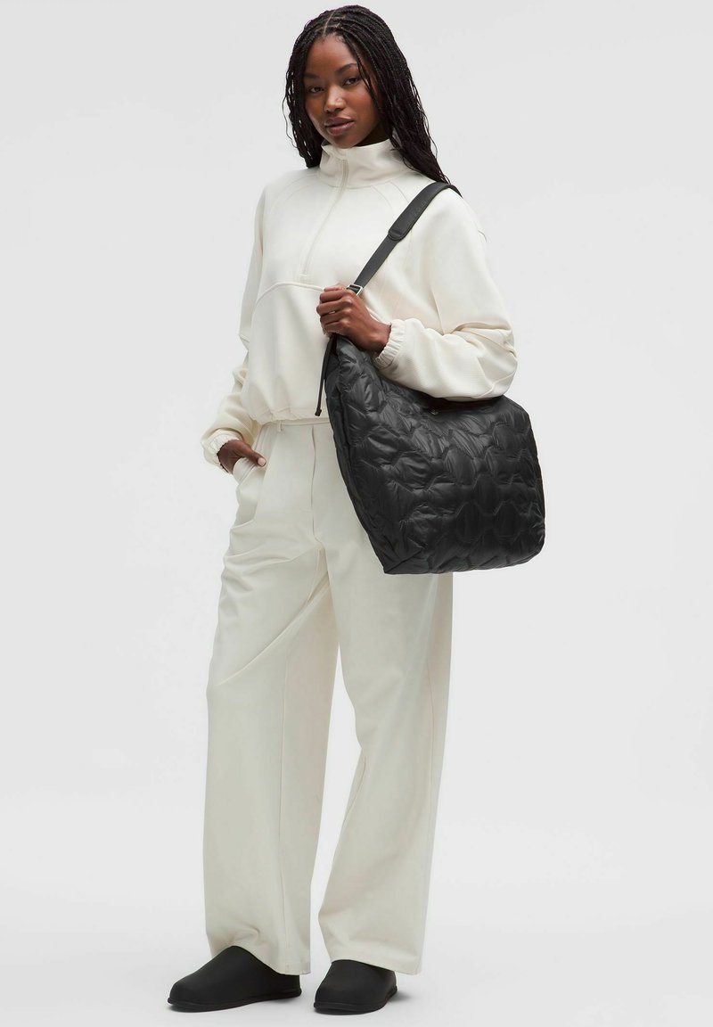 Czarna pikowana torebka na ramię z teksturowaną powierzchnią i regulowanym paskiem. Modelka ma na sobie kremową bluzę i luźne białe spodnie.