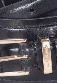 Närbild av ett svart läderbälte med ett guldtonat spänne och en rektangulär metallöga i guld, graverad med varumärke.