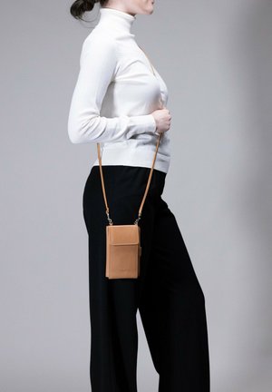 Tan leather crossbody phone pouch with a front flap, adjustable strap, and smooth texture, worn by a person in a white turtleneck and black pants.