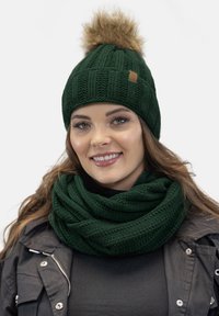 Dark green knitted beanie with a faux fur pom-pom on top and a matching green textured scarf, displaying ribbed and woven patterns.