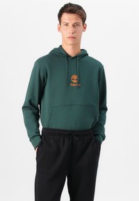 Grønn hettegenser med kengurulomme, med et orange Timberland-logo. Kombinert med svarte joggebukser med elastisk liv. Glatt tekstur.