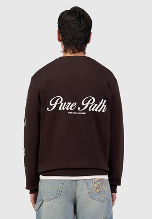 Man with dark hair wearing a dark brown sweatshirt with "Pure Path Own Your Journey" text on back and light blue jeans with embroidered patch.