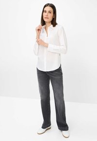 Weißes Hemd mit spitzem Kragen, langen Ärmeln; kombiniert mit dunkelgrauen, weit geschnittenen Jeans und weißen Sneakers. Glatte Stofftextur.