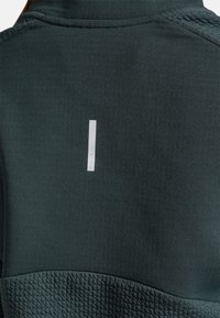 Mørkegrøn jakke med en glat overflade og en ribbet nederste panel, med et hvidt rektangulært mærke, der står "X-LITE" på ryggen.