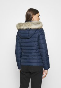 Chaqueta de plumón de color azul marino con capucha con borde de piel sintética, patrón de acolchado horizontal y puños elásticos. Vista trasera que muestra un diseño ajustado.