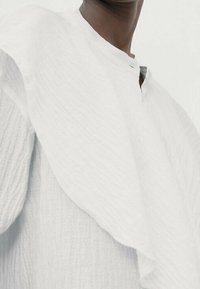 Blouse blanche texturée avec un design à volants superposés à l'épaule et une petite patte de boutonnage à l'avant avec un décolleté subtil.