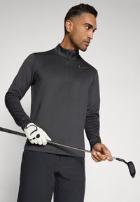 Tamno sivi pulover dugih rukava s polu-zatvaračem i zlatnim Nike logom. Muškarac u bijelom golf rukavicu drži golf štap.