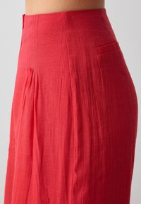 Jupe rouge taille haute en tissu texturé. Présente des plis et une poche dissimulée. Design lisse et fluide adapté à diverses occasions.