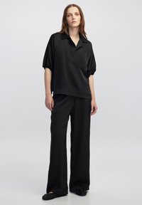 Blusa nera a maniche corte con colletto e taschino sul petto, abbinata a pantaloni neri a vestibilità comoda e scarpe slip-on decorate.