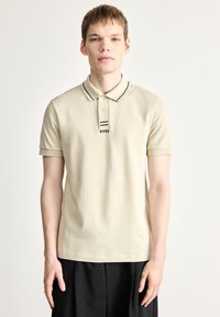 Μπεζ πόλο πουκάμισο με μαύρο ριμπ γιακά και μανσέτες στα μανίκια. Διαθέτει μικρό μαύρο λογότυπο "BOSS" στο στήθος. Μαλακό βαμβακερό ύφασμα.