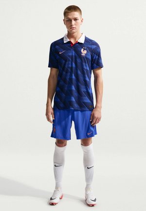 Jonge mannelijke atleet staat in het voetbaltenue van Frankrijk, met een blauw gemêleerd shirt, blauwe korte broek, witte sokken en voetbalschoenen tegen een witte achtergrond.