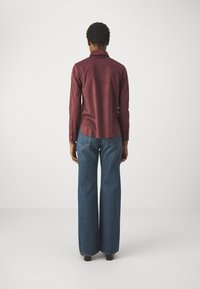 Bordowa koszula z długim rękawem wykonana z gładkiej tkaniny, zestawiona z niebieskimi dżinsami o wysokim stanie i szerokich nogawkach. Posiada klasyczny kołnierzyk i mankiety zapinane na guziki.
