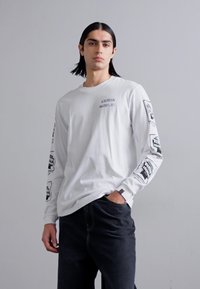 White long-sleeve shirt with black graphic prints on sleeves, paired with dark denim jeans, featuring a relaxed fit and simple design.