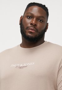 Bež majica od pamuka kratkih rukava, s bijelim tiskanim natpisom "JACK & JONES Originals Studio" na prednjem dijelu. Glatka tekstura.