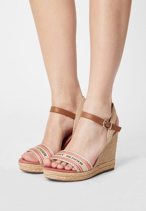 Pieds portant des sandales compensées beiges en tissu tressé avec des brides marron à la cheville et des brides en tissu de marque sur les orteils, sur fond blanc.