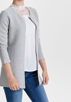 Femme portant un cardigan gris clair ouvert sur un haut en dentelle blanche et un jean bleu, debout, les mains détendues le long du corps.