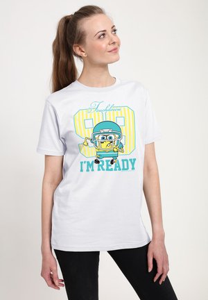 Ung kvinde iført hvid T-shirt med tegneserie-fodboldspiller og teksten "Touchdown 98 I'm Ready," der kigger op og smiler.
