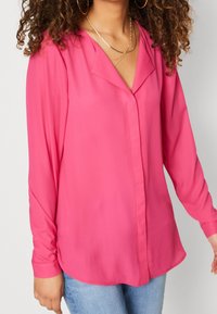 Blouse rose à manches longues avec un col en V et des boutons sur le devant, fabriquée en tissu lisse, présentant un ourlet incurvé et une coupe décontractée.