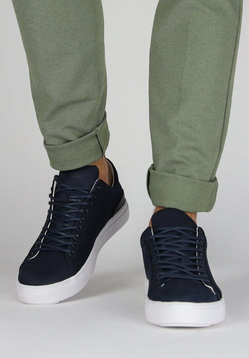 Marineblauwe sneakers met een gladde textuur, voorzien van een laag ontwerp, witte rubberen zolen en vetersluiting. Gecombineerd met opgerolde olijfgroene broek.