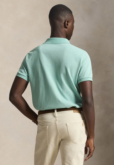 Άνδρας που φοράει κοντομάνικο πόλο σε μέντα πράσινο, κλεισμένο μέσα σε εκρού παντελόνι με καφέ ζώνη, εμφανιζόμενος από πίσω σε μπεζ φόντο.