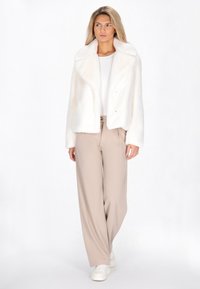 Giacca in pelliccia sintetica bianca con colletto ampio, chiusa con due bottoni, abbinata a pantaloni a gamba larga beige chiaro e sneakers bianche.