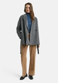 Grijze wolblendjas met een losse, oversized pasvorm, voorzien van een enkele knoopsluiting, gedragen over een blauwe denimshirt en beige broek.