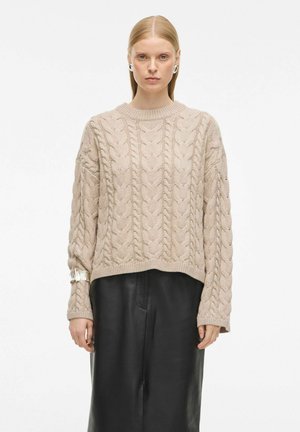 Beige gebreide trui met kabelpatroon, ronde halslijn en verlaagde schouders, gecombineerd met een zwarte leren rok. Zilveren hardware-accent.