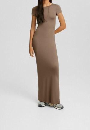 Femme portant une robe longue marron cintrée à manches courtes, associée à des baskets grises et beiges sur un fond uni.