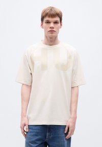 Μπεζ κοντ ομάνικη μπλούζα με μεγάλο ανοιχτόχρωμο γραφικό "HU" μπροστά. Μαλακό βαμβακερό ύφασμα και άνετη εφαρμογή. Φορεμένη με μπλε τζιν.