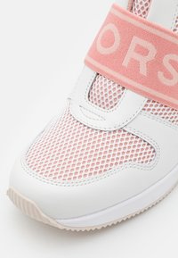Bout de basket blanc et rose avec tissu mesh, cuir lisse, semelle beige et large sangle élastique rose sur le dessus avec lettrage partiel.