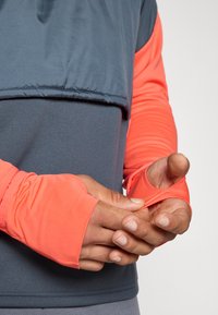 Langærmet atletisk skjorte med et koralfarvet overlag og et mørkeblåt underlag. Ærmerne har tommelhuller for ekstra dækning.