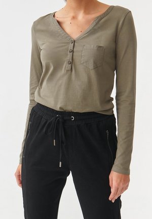 Personne portant une chemise henley à manches longues vert olive avec une poche avant et un pantalon noir à cordon de serrage avec des poches zippées.