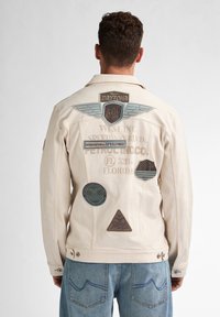 Chaqueta de mezclilla beige con varios parches bordados, incluyendo 'DAYTONA 500' y diseños circulares, con cierres de botones y un ajuste relajado.