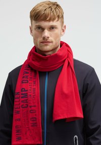 Young man with short blond hair wearing a navy jacket and a red scarf with black printed text, facing forward against a plain background.
