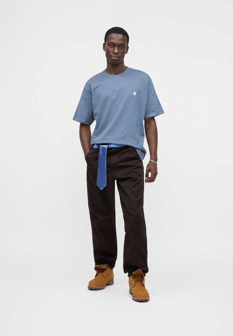 Uomo in piedi con le mani nelle tasche, indossa una maglietta blu, pantaloni neri con cintura blu e stivali da lavoro marroni su sfondo bianco.