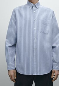 Jasnoniebieska koszula z długim rękawem, zapinana na guziki, z białymi pionowymi paskami, z kołnierzykiem w kształcie ostrego oraz z kieszenią na klatce piersiowej po lewej stronie.