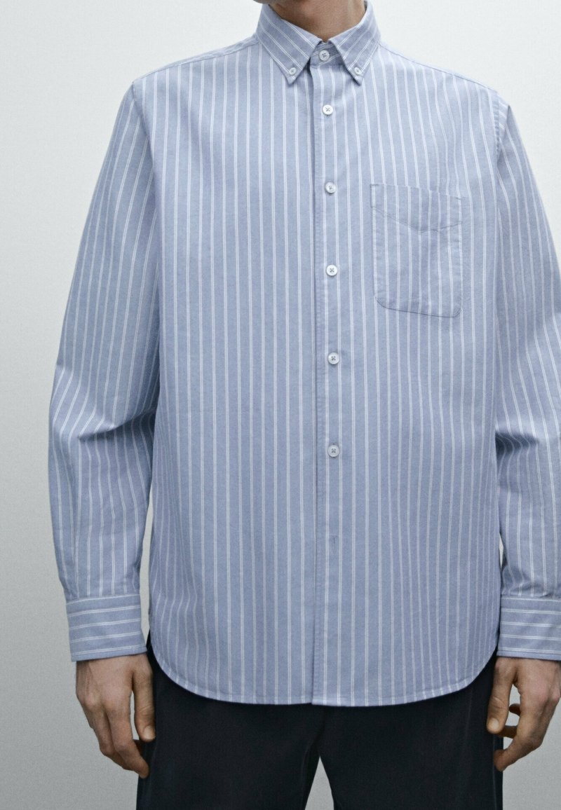Jasnoniebieska koszula z długim rękawem, zapinana na guziki, z białymi pionowymi paskami, z kołnierzykiem w kształcie ostrego oraz z kieszenią na klatce piersiowej po lewej stronie.