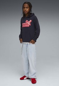 Sweat à capuche bleu marine avec grand logo "PUMA" rouge, associé à un jean large bleu clair et des baskets rouges. Design simple, tissu en coton, coupe décontractée.