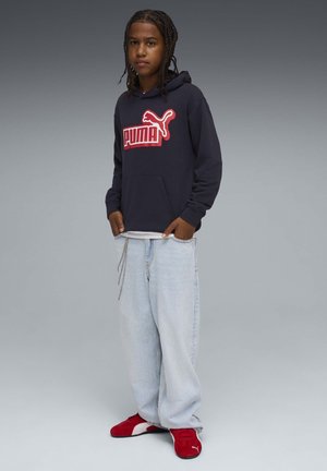 Felpa con cappuccio blu navy con grande logo rosso "PUMA", abbinata a jeans larghi azzurri e sneaker rosse. Design semplice, tessuto in cotone, vestibilità casual.