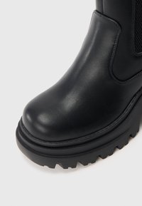 Μαύρη δερμάτινη μπότα με χοντρή σόλα με ανάγλυφα και ελαστική πλαϊνή φάσα, σε κοντινή λήψη σε λευκό φόντο.